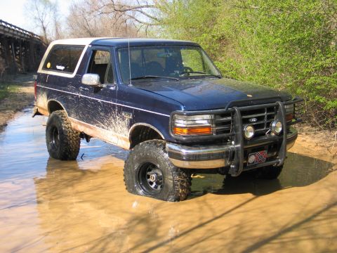 1995 Ford Bronco - A muddy blue Ford Bronco with a white top navigates through a water-filled dirt path.
