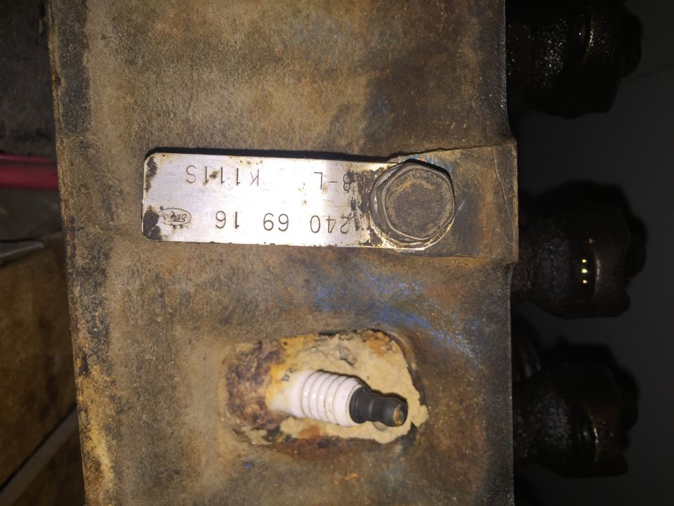 Close-up of a rusty engine component with a metal tag showing numbers and letters, and a spark plug. The tag has 'Ford' logo
