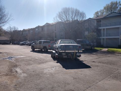 1987 Ford Crown Victoria LTD - Pickup truck tows a 1987 Ford Crown Victoria LTD on a trailer in front of apartment buildings.