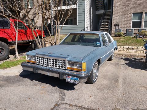 1987 Ford Crown Victoria LTD - This 1987 Ford Crown Victoria LTD is seen from the front-right in a parking lot. A red off-road vehicle is parked to its left.
