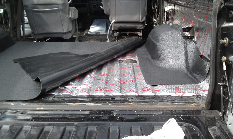 Vehicle interior with sound deadening material on the floor and insulation on the walls. A black plastic trim piece is visible