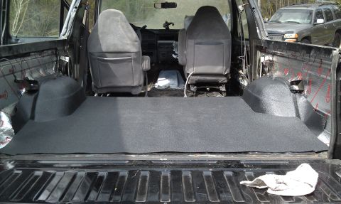 1995 Ford Bronco - The interior of a vehicle shows newly installed dark grey flooring in the cargo area, with front seats visible. A second vehicle is parked…