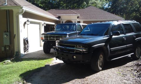 1995 Ford Bronco - Two SUVs, a lifted Ford Bronco and a dark Chevrolet Tahoe, are parked in a driveway in front of a house.