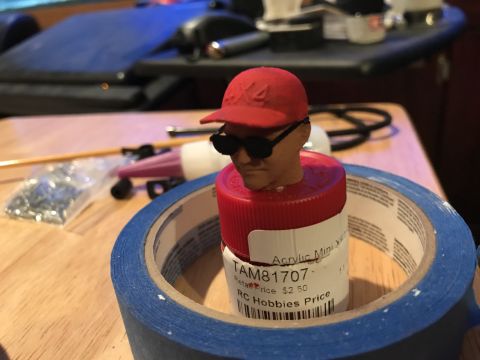 1989 Ford Bronco - A model figure head wearing a red cap and sunglasses is placed on a bottle cap, surrounded by blue tape and hobby supplies.