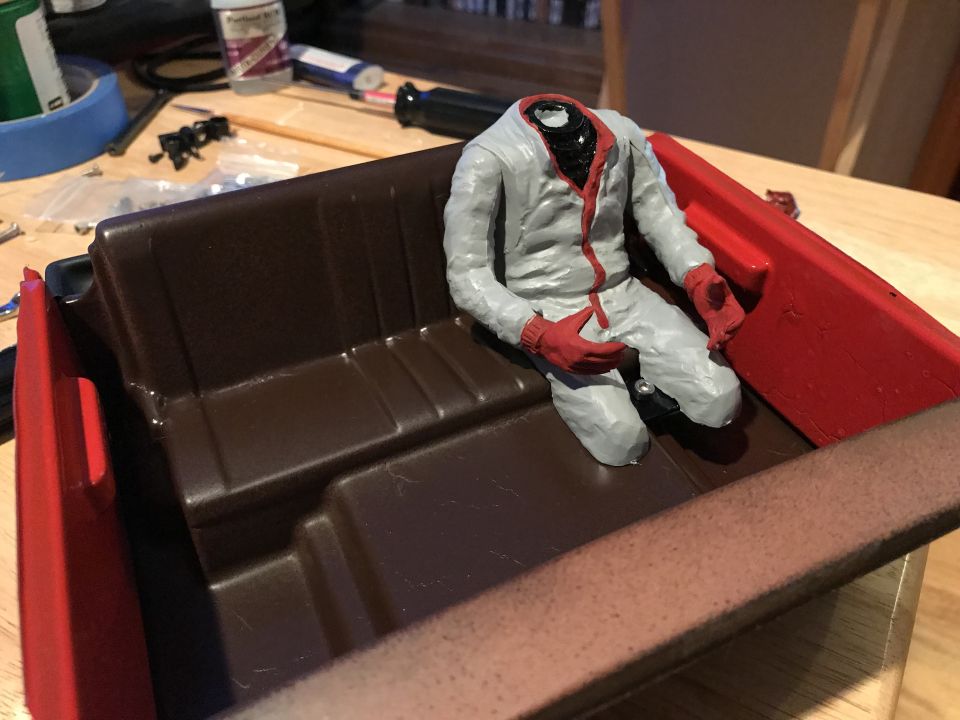 Model car interior with brown seats and red trim, a headless figure in a gray jumpsuit with red gloves