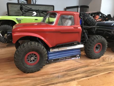 1989 Ford Bronco - A red RC truck body is propped up by a stack of DVDs, with other RC vehicles in the background.