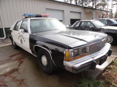 1988 Ford Crown Victoria LTD - A two-tone police car with a light bar is parked outside a building, with another patrol car visible behind it.