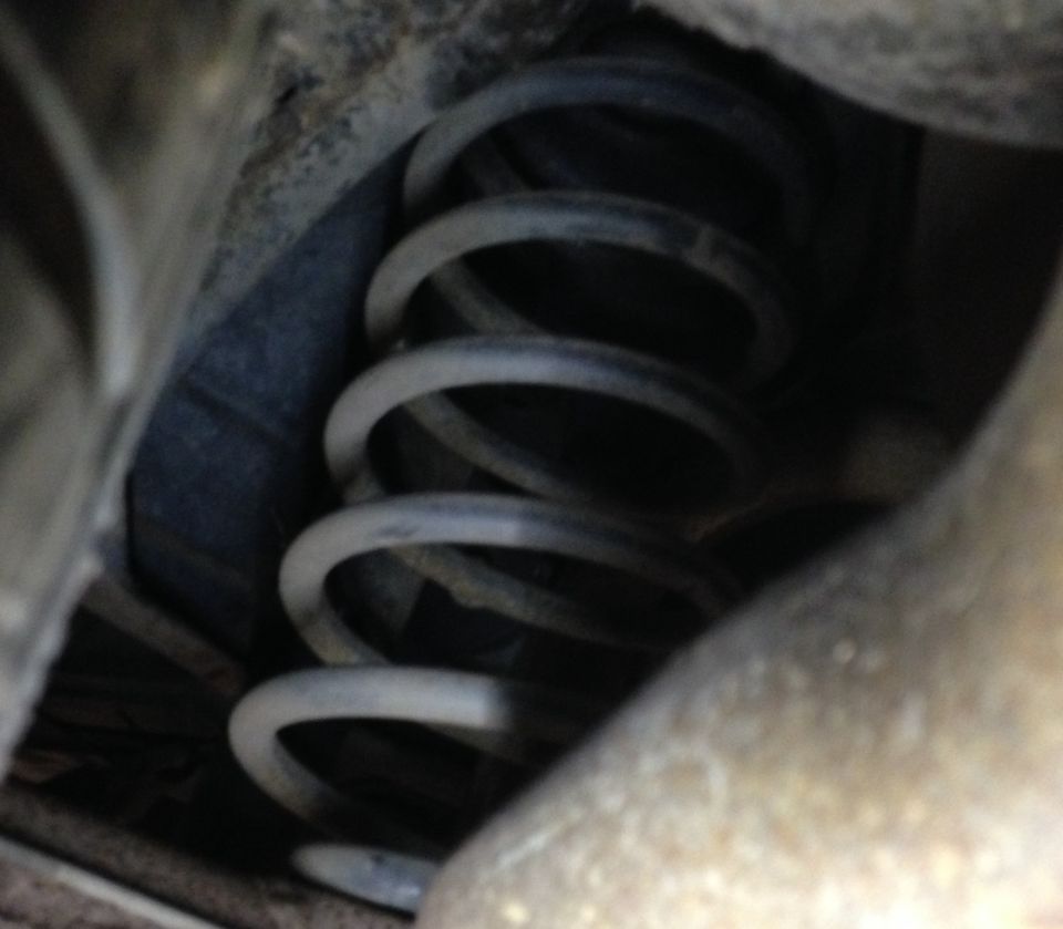 Coil spring in a dark, dirty vehicle suspension system. Text overlay reads SUPERMOTORS.NET