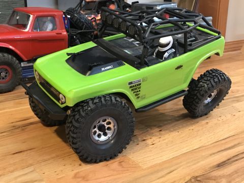 1989 Ford Bronco - A green remote-controlled car with a roll cage and driver is parked on a wooden floor next to a red truck.