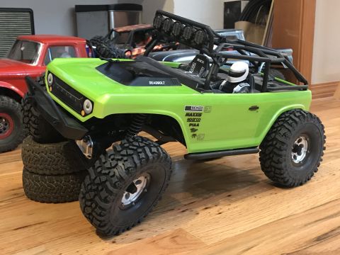 1989 Ford Bronco - A lime green RC rock crawler with a roll cage and driver figure is parked on a wooden floor, with other RC vehicles in the background.