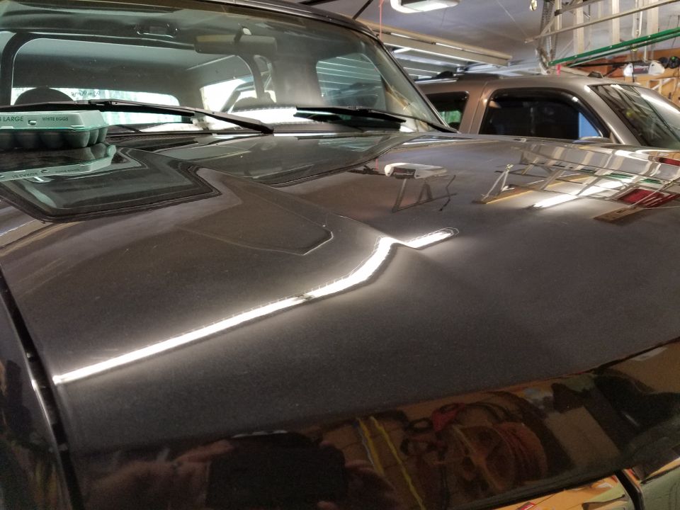 Dark SUV hood with a dent, windshield wipers, and an egg carton. Another SUV is visible behind it