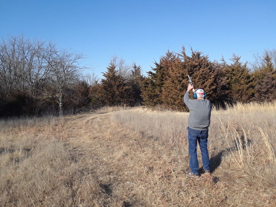man shooting a rifle in a dry field with trees and blue sky