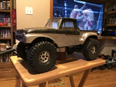 1989 Ford Bronco - RC rock crawler truck with large tires sits on a wooden table in front of a television.