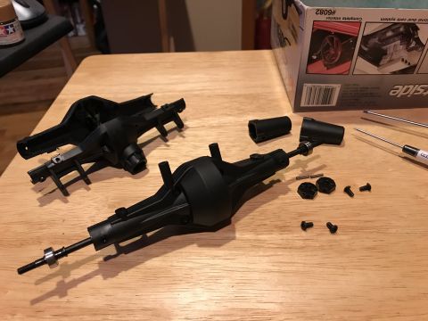 1989 Ford Bronco - Two black plastic RC car axle housings are laid out on a wooden surface with various small parts including screws, pins, and bearings. A pa…