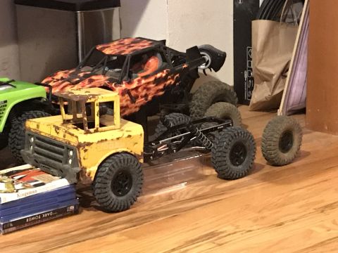 1989 Ford Bronco - A yellow, weathered toy truck sits on a stack of books in front of a black and orange toy monster truck. Two loose toy truck tires are visi…