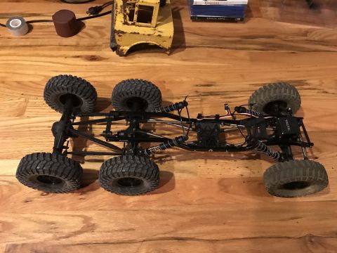 1989 Ford Bronco - The chassis and six wheels of a remote-controlled rock crawler are shown from a top-down perspective on a wooden surface. The vehicle has a…