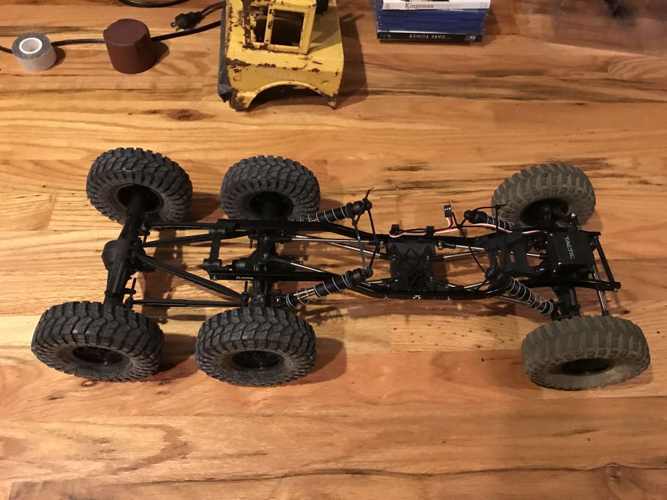 Remote-controlled rock crawler chassis with six wheels on a wooden floor.