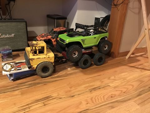 1989 Ford Bronco - A yellow toy forklift is parked on a wooden floor, carrying a stack of books. Behind it, a green remote-controlled off-road vehicle is on t…