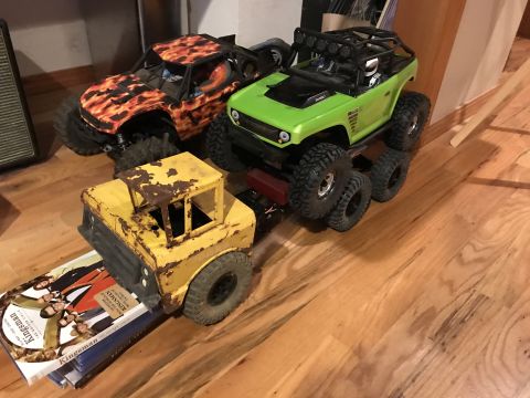 1989 Ford Bronco - Three remote-controlled toy trucks are displayed on a wooden floor. A yellow truck with a rusty appearance is in the foreground, followed b…