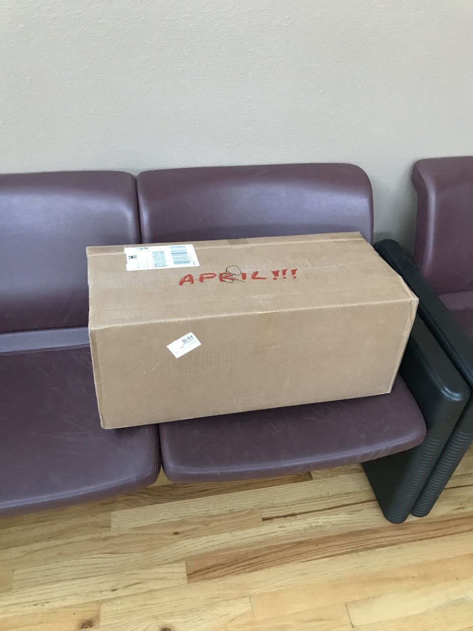Cardboard box with "APRIL!!!" written in red marker on burgundy chairs.