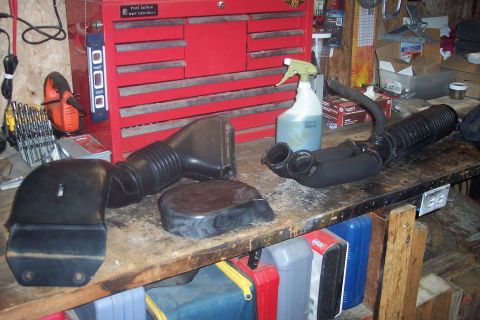 1995 Ford Bronco - Two black plastic intake components are laid out on a wooden workbench in front of a red metal toolbox. A spray bottle and other tools are…