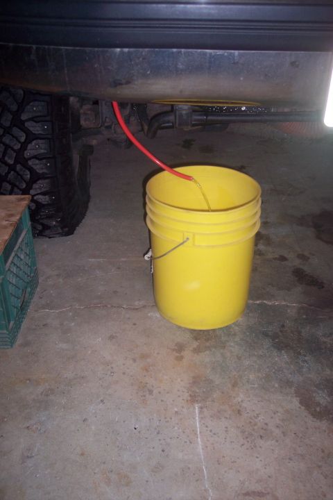 1995 Ford Bronco - A red hose drains liquid from under a vehicle into a yellow bucket. The vehicle's tire and part of its undercarriage are visible.