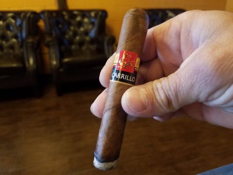 2018 Ford Bronco - A hand holds a cigar with a red and gold band that reads "CARRILLO". In the blurred background, two dark leather armchairs are visible.