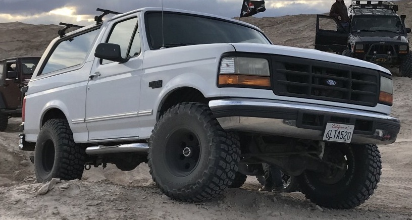 White 1990s Ford Bronco with off-road tires parked on sandy terrain, black Jeep in background.
