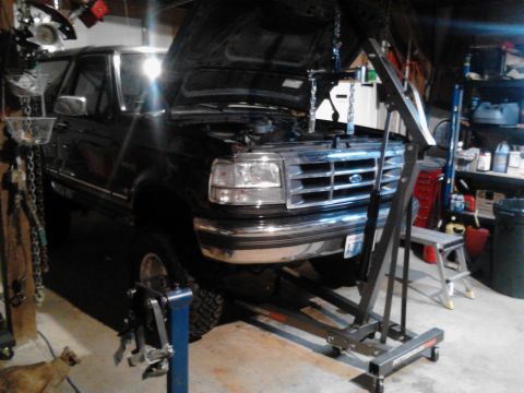 1995 Ford Bronco - A black Ford Bronco is parked in a garage with its hood open, supported by an engine hoist. The vehicle is on a lift, and tools and shelves…