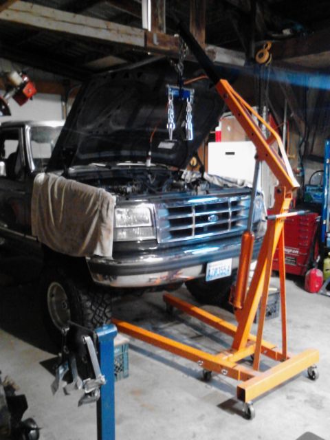 1995 Ford Bronco - An orange engine hoist is positioned in front of a dark-colored pickup truck with its hood open. The truck appears to be undergoing mainten…