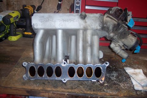 1995 Ford Bronco - A silver aluminum intake manifold with a throttle body attached is displayed on a wooden workbench. The manifold has the part number RF-E8T…