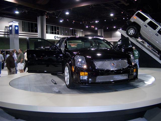 Black Cadillac sedan with doors open on a display turntable, a silver Hummer on a ramp behind it