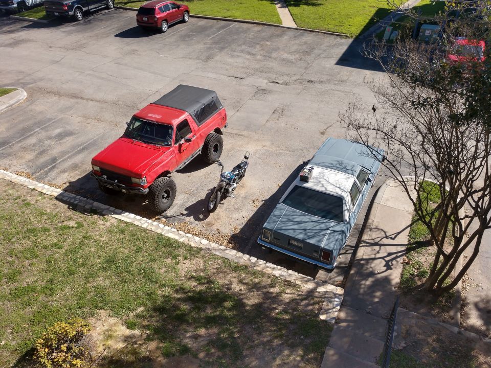 Red lifted Ford Bronco and blue/white sedan parked in lot with motorcycle.