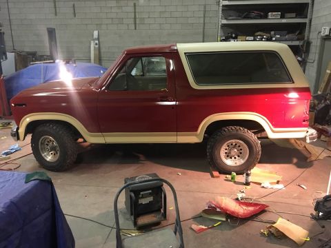1985 Ford Bronco - A red and tan 1985 Ford Bronco is parked in a garage with parts scattered on the floor around it. The vehicle appears to be in the process…