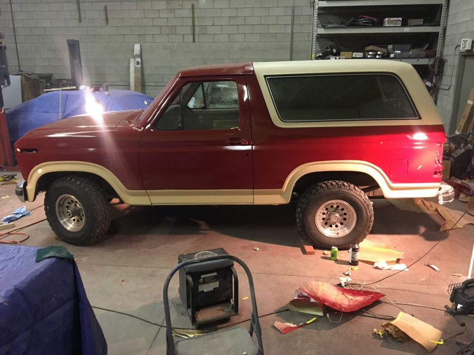 Red and tan 1985 Ford Bronco in a garage, undergoing restoration.