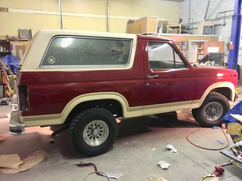 1985 Ford Bronco - The side profile of a red and tan Ford Bronco is shown in a workshop setting. The vehicle appears to be undergoing some form of restoration…