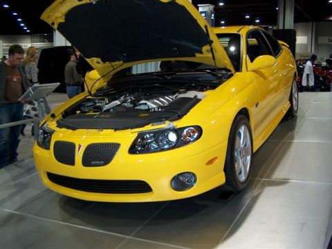 2001 Ford Crown Victoria - Yellow Pontiac GTO displayed at an auto show, hood raised to reveal the engine.