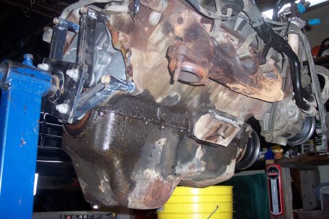 1995 Ford Bronco - The underside of a V8 engine, likely from a 1995 Ford Bronco, is shown mounted on an engine stand. The oil pan is heavily soiled with oil a…