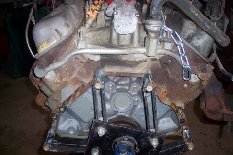 1995 Ford Bronco - A close-up, slightly angled view of a V8 engine block, heavily rusted in some areas and painted gray in others. A metal chain is attached t…
