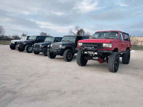 1989 Ford Bronco - A line of off-road vehicles, including a red SUV and multiple Jeeps, are parked on a gravel lot under a cloudy sky.