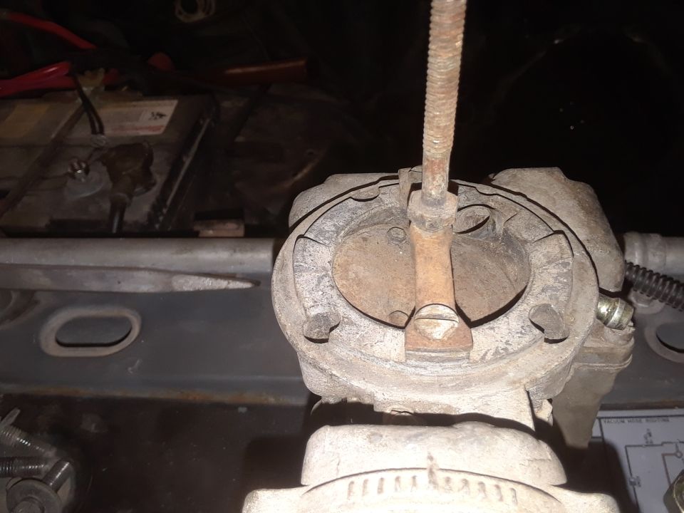 Close-up of a rusty carburetor with a metal rod, showing engine parts and a battery in the background