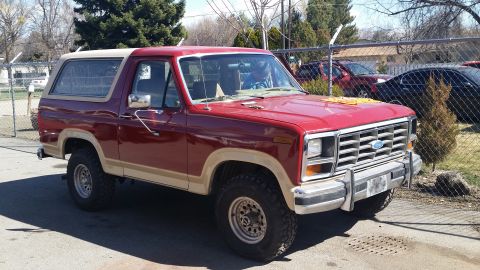 1985 Ford Bronco - This red Ford Bronco with tan trim is parked outdoors, with visible rust repair on its rear quarter panel.