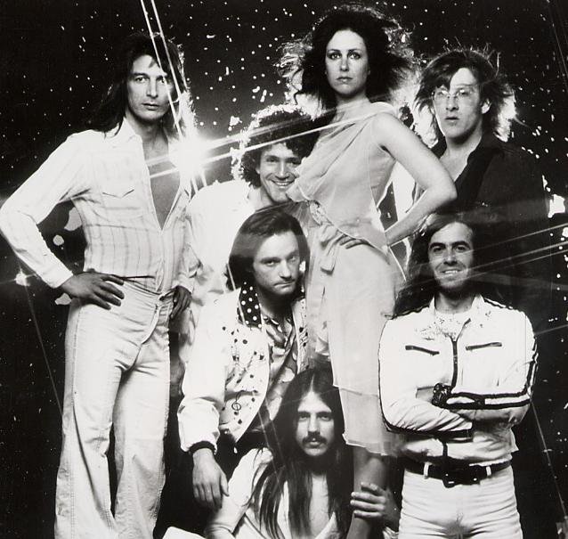 Seven people in 70s attire pose against a starry background. One person in front has long hair and a mustache