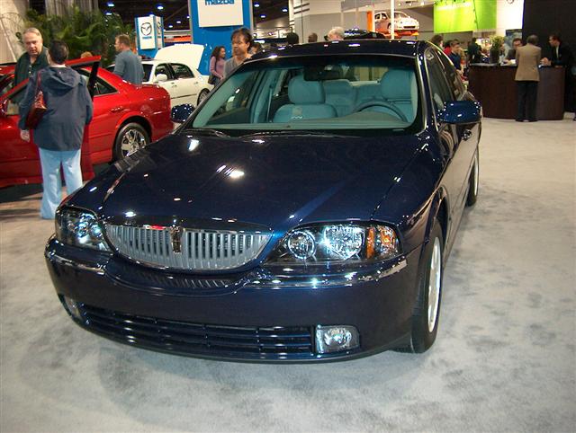 Dark blue Lincoln LS sedan with chrome grille at an auto show. Red car and white car visible behind it