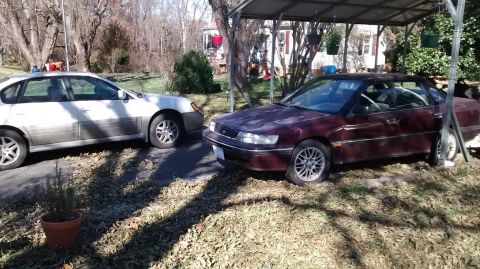 1994 Subaru Legacy - A maroon 1994 Subaru Legacy sedan is parked next to a white sedan under a carport.
