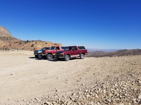 1967 Ford Bronco - Several Ford Broncos and trucks are parked on a dusty, rocky terrain under a clear blue sky.
