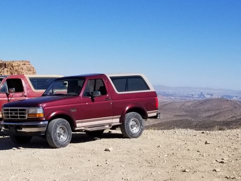 1967 Ford Bronco - This maroon Ford Bronco with a tan top is parked on a desert overlook, with another vehicle partially visible behind it.
