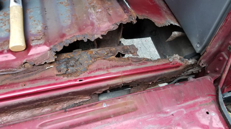Severely rusted floor pan and rocker panel of a red vehicle