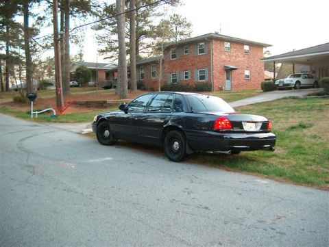 2001 Ford Crown Victoria - A dark-colored sedan is parked on the side of a road in front of a brick building.