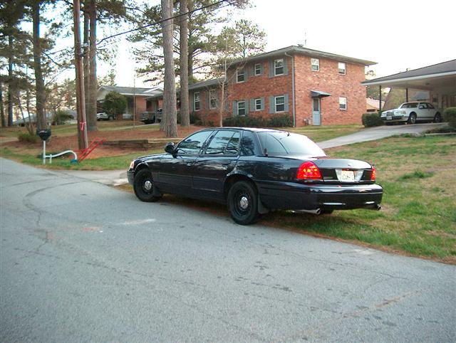 Black Ford Crown Victoria parked on a residential street. Another white car is visible under a carport